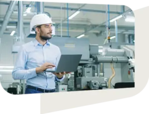 A professional man wearing a white hard hat and light blue shirt is standing in an industrial facility, holding a laptop