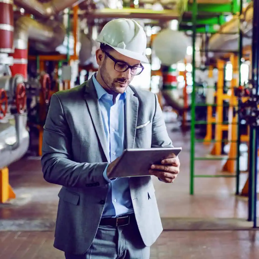 interim manager in gray suit and with helmet on head using tablet while standing in power plant
