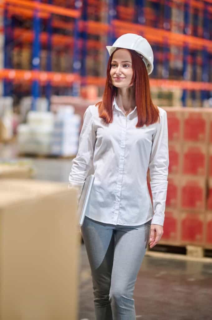 Smiling young woman in white blouse and safety helmet with laptop standing looking to side at large lighted warehouse