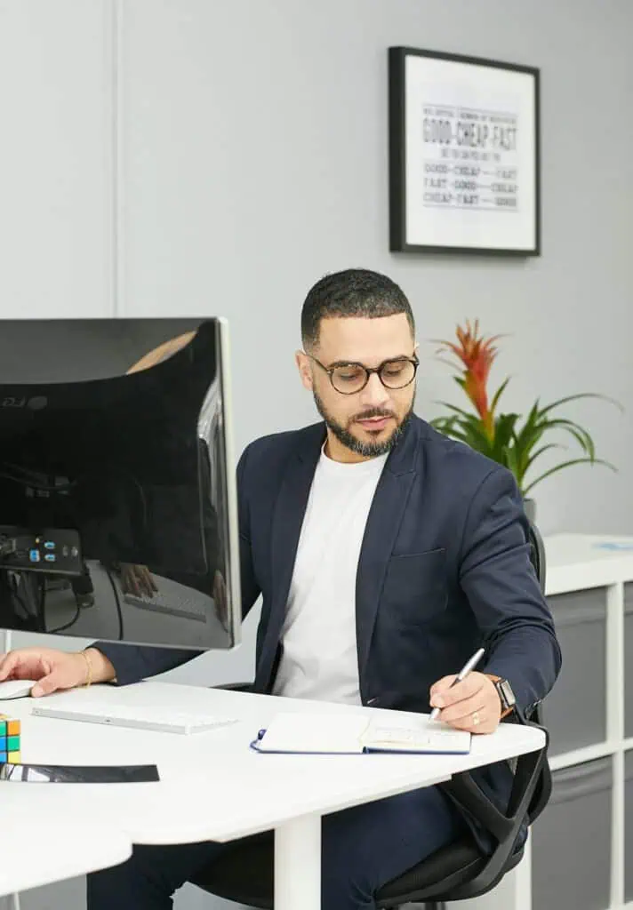 Operations manager in a suit sitting at is desk behind computer, taking notes in his notepad