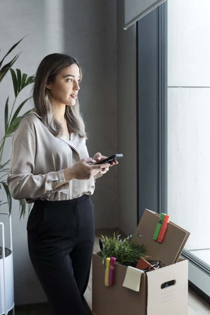 Relocation manager looking out of window with phone in her hands and box with her stuff sitting on the window ledge