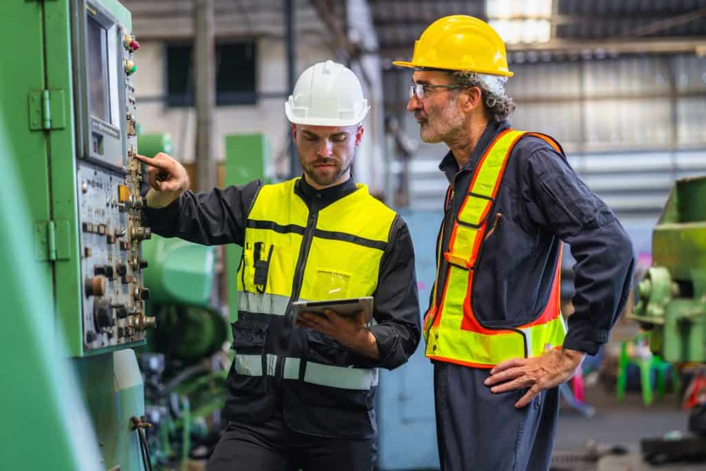 Two men in safety gear are standing next to a machine, one pointing at a button, they are discussing safety