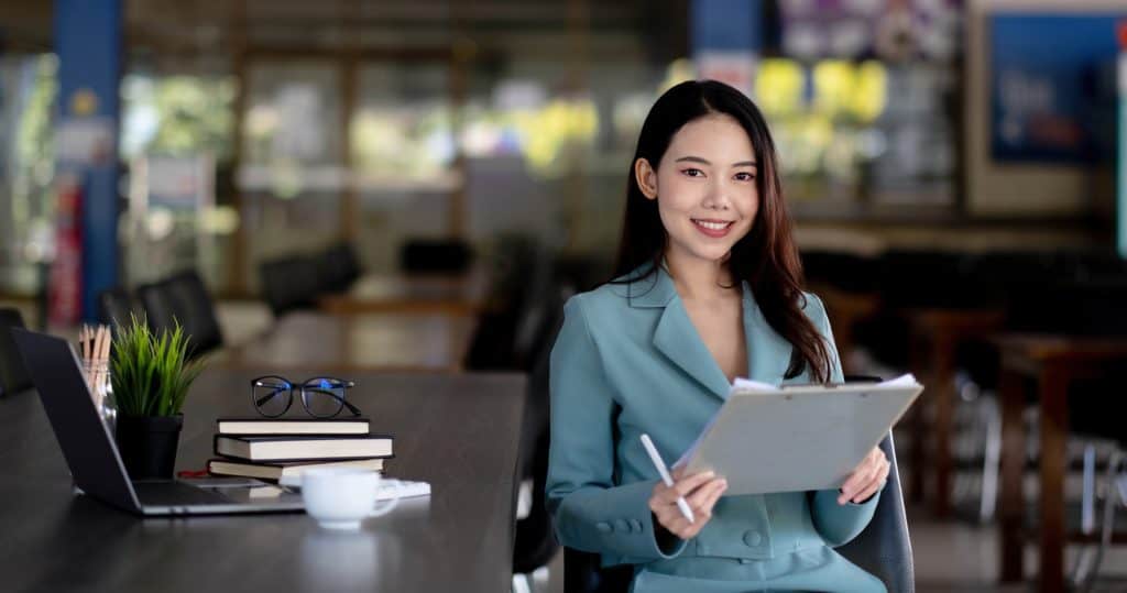 Smiling young asian business woman is sitting in office with papers in hands