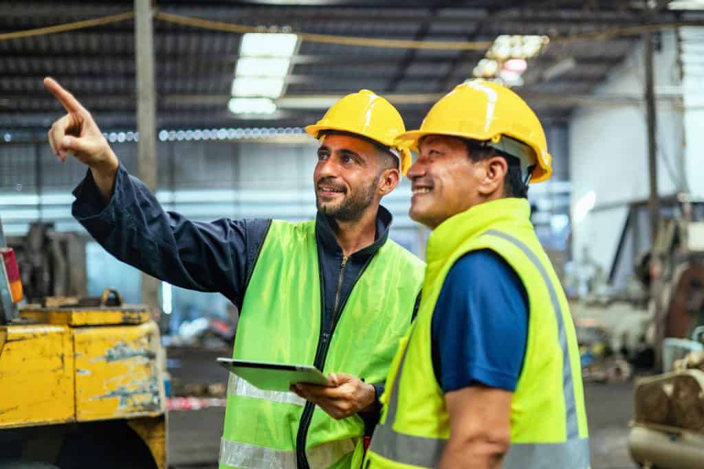 Two men in safety vests are pointing at something on a tablet. They are smiling and seem to be enjoying their work