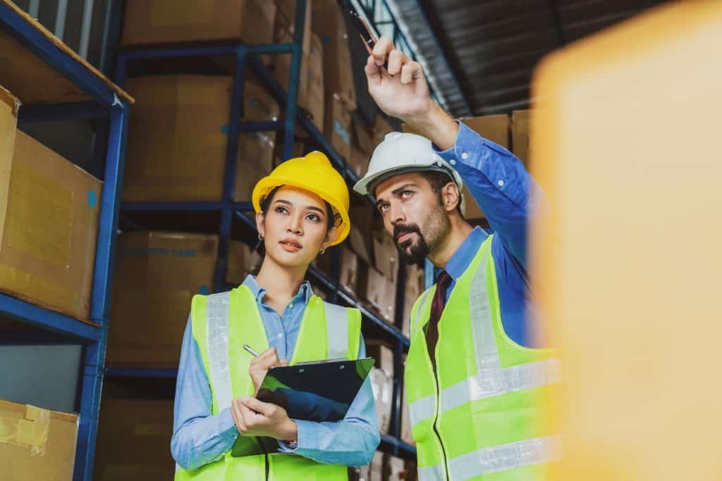 Two logistic experts wearing reflexive vest and hard helmets stand in warehouse, with the man pointing out and the woman taking notes