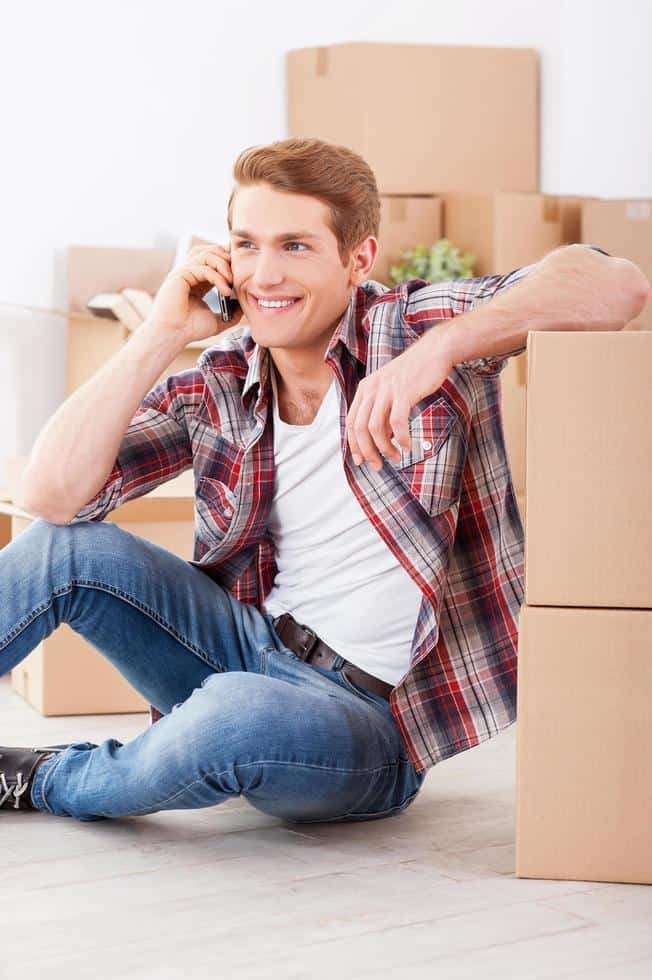 Young man sitting on the floor and talking on the mobile phone with cardboard boxes around him