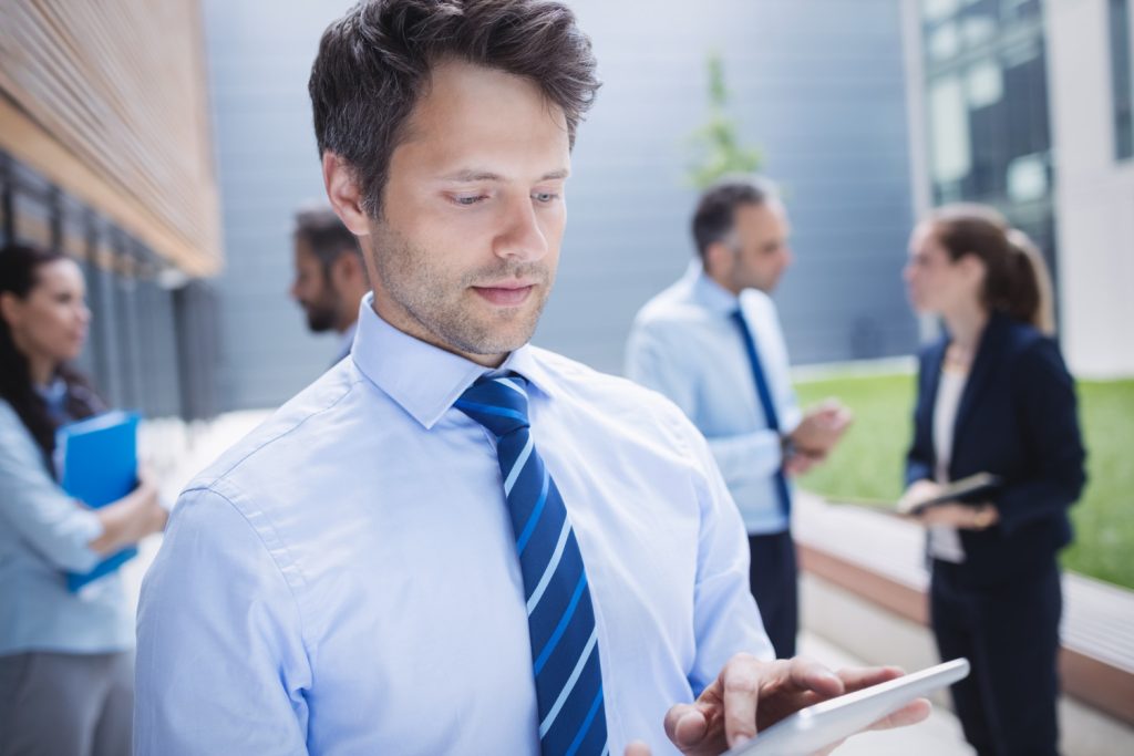 Sales executive search expert in blue shirt and tie holds tablet in his hand, scrolling. He's standing outside a building with talking business people in the backround.
