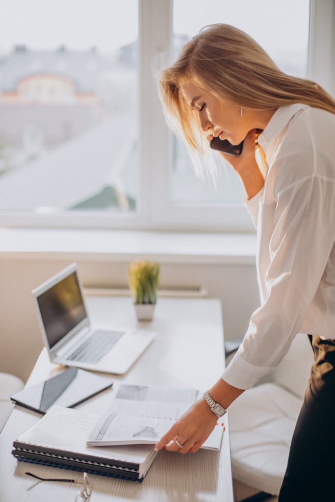 Female sales executive search expert is standing over her desk in office, talking on a phone with papers in the other hand